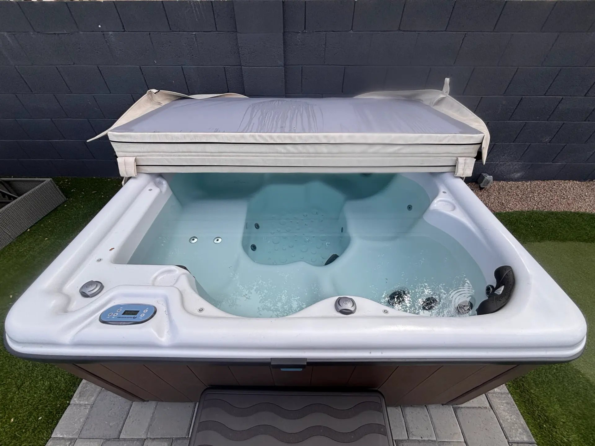 Private hot tub with the cover folded back, beside turf and a dark block wall