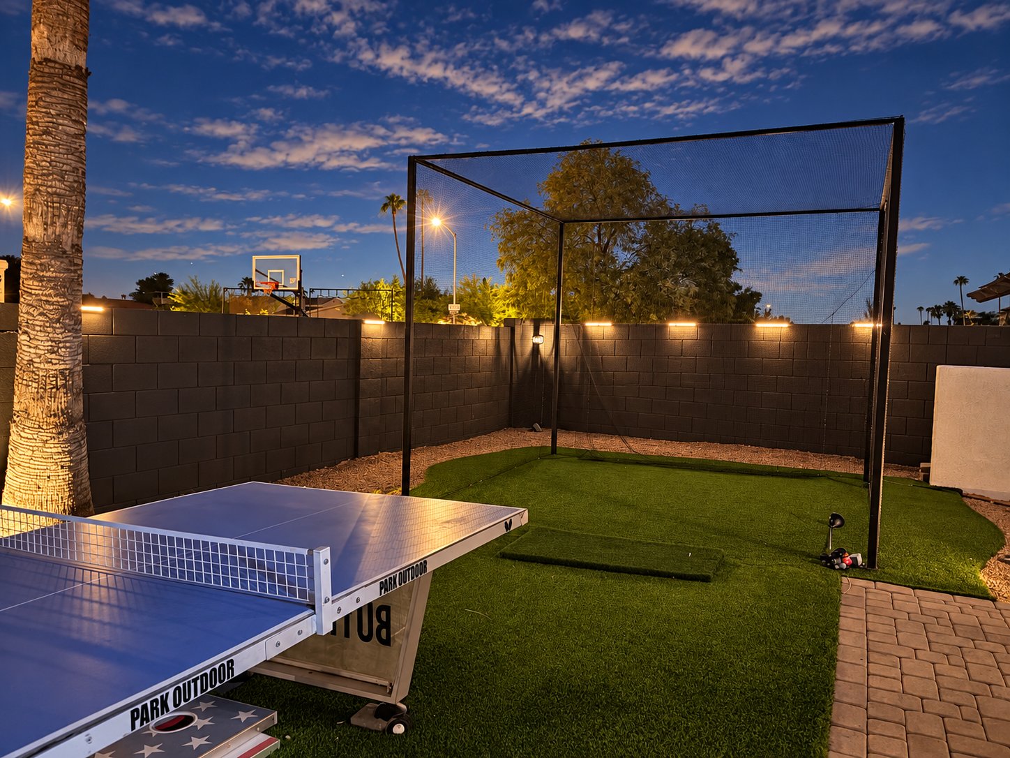 Netted golf hitting cage with turf hitting mat at dusk, outdoor ping-pong table in front